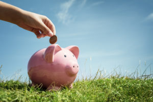 Child hand inserting coin and saving money in piggy bank with grass and blue sky background